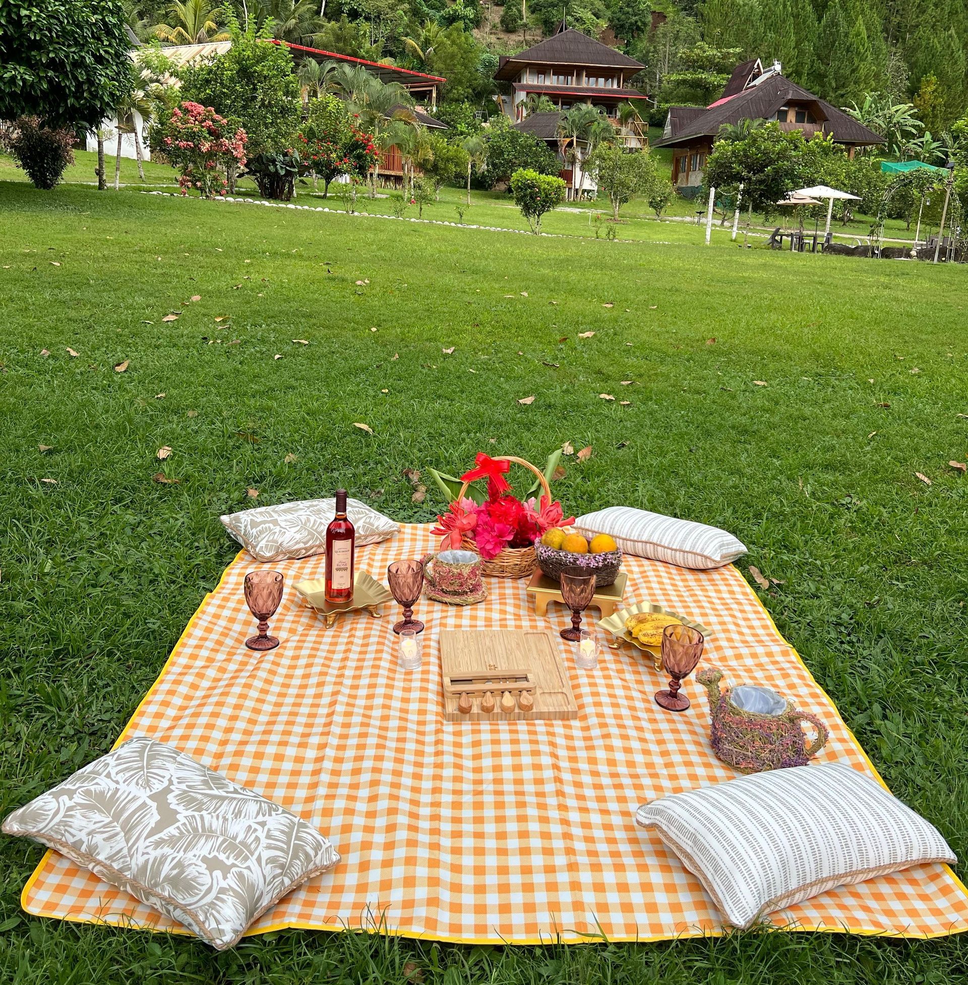 Un picnic preparado sobre una manta a cuadros amarilla con comida y vino en un césped de un jardín.