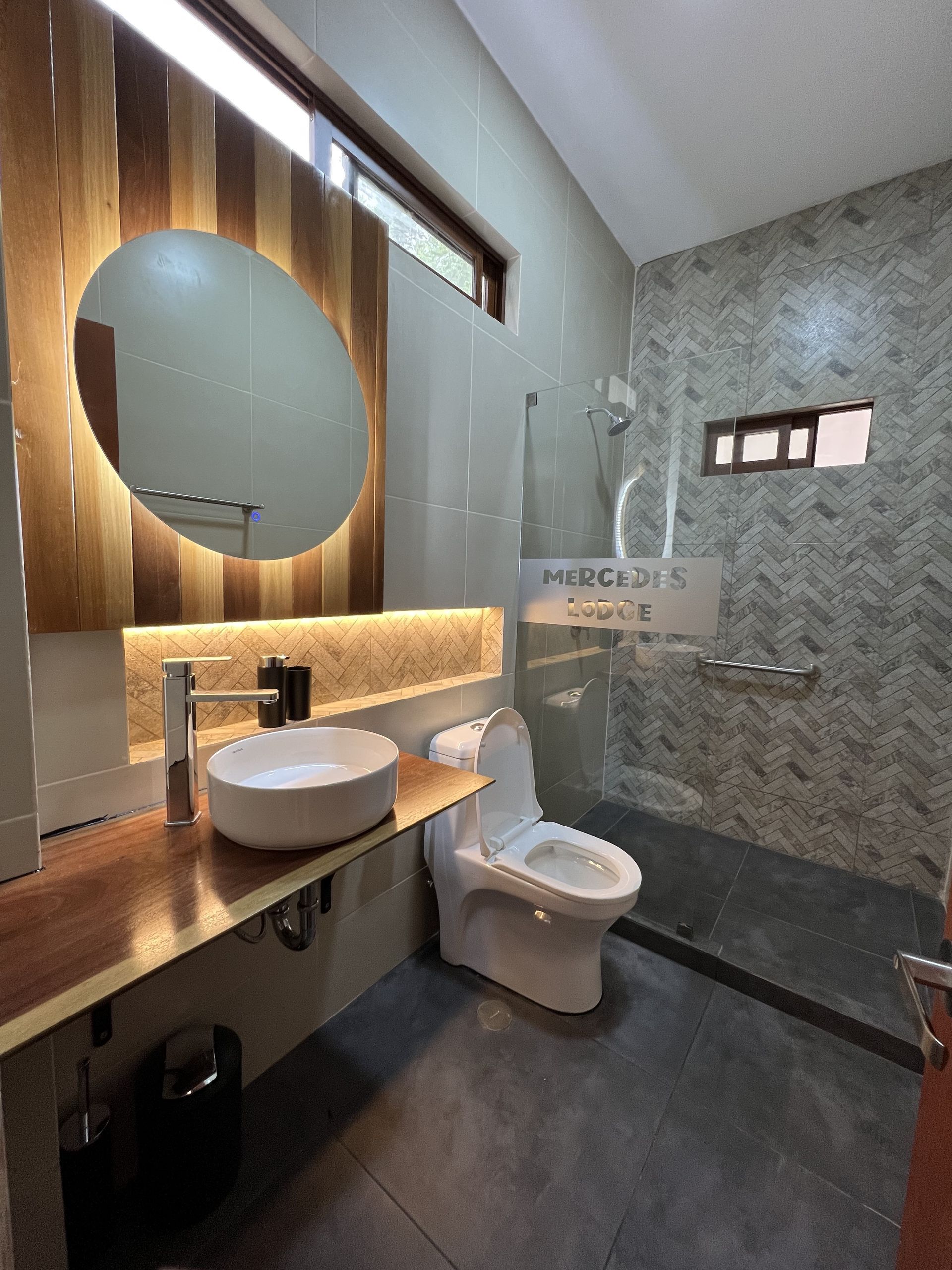 Baño moderno con espejo redondo, lavabo sobre encimera, inodoro y zona de ducha alicatada.