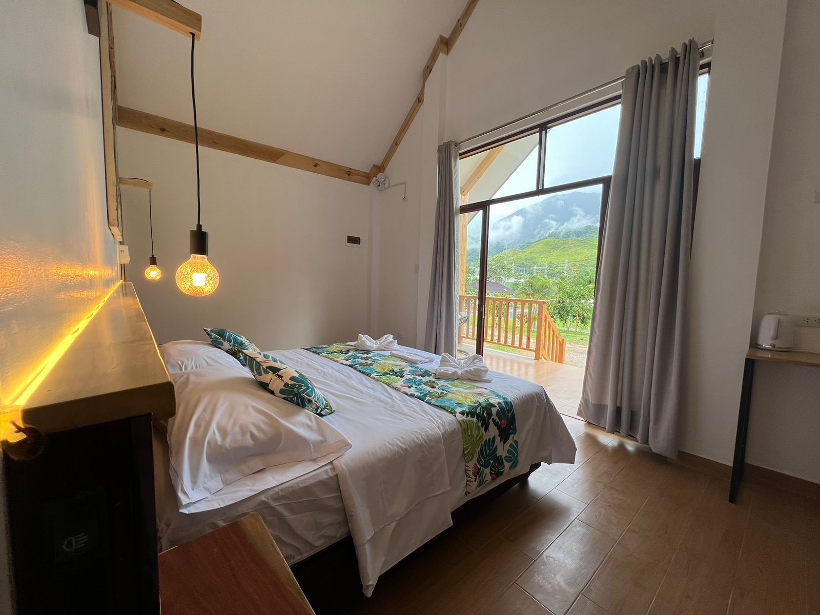 Habitación luminosa con cama hecha, lámparas colgantes y puertas correderas abiertas con vistas a la montaña.