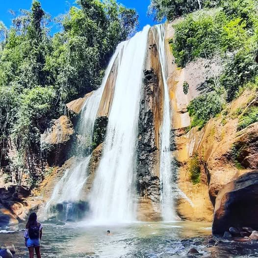 Una alta cascada cae por acantilados rocosos hasta formar una poza, con una persona de pie en primer plano.