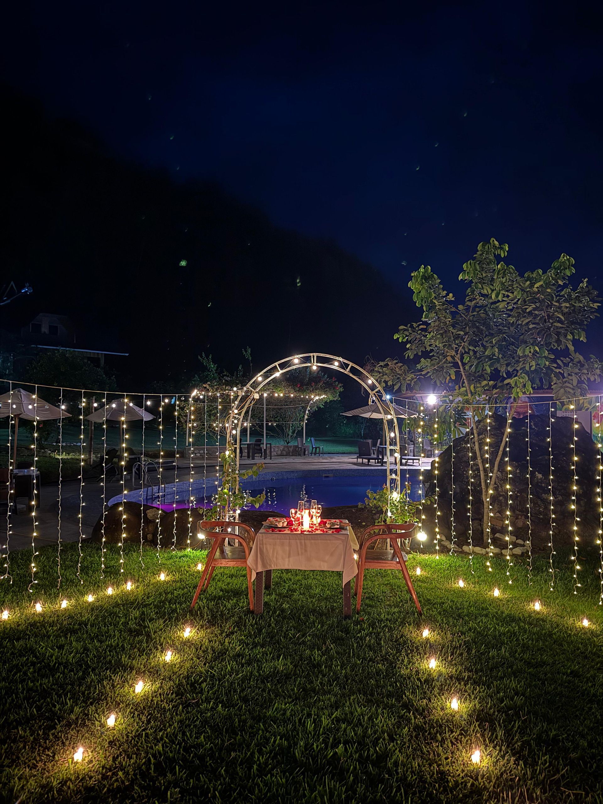 Sendero iluminado en el jardín con guirnaldas de luces, arco y una mesa con vistas al agua por la noche.