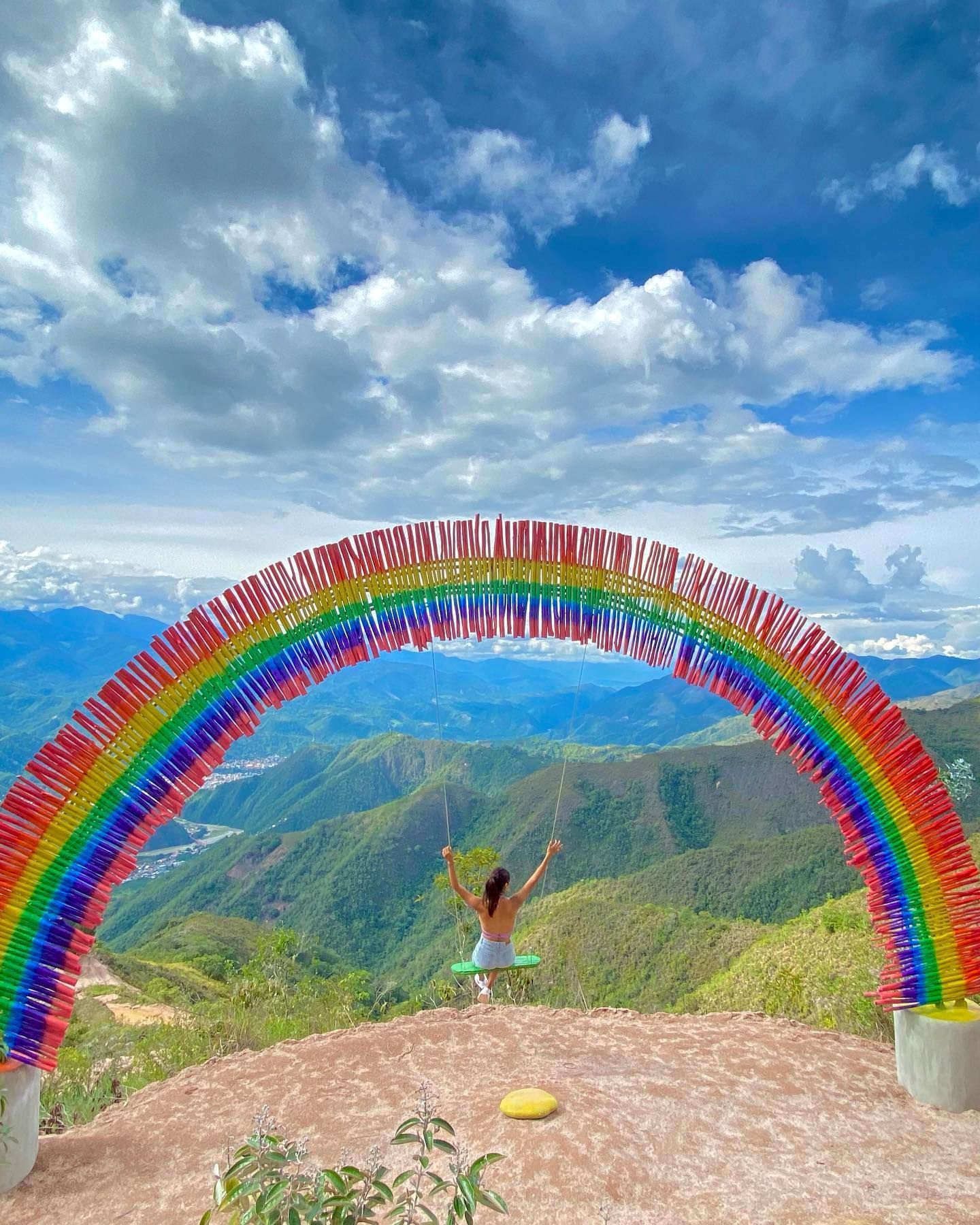 Una persona se encuentra bajo un arcoíris, contemplando montañas verdes y un cielo azul nublado.