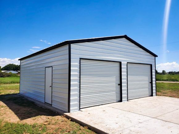 A white metal two-car garage with black trim, a side entrance door, and a concrete driveway in a rural field setting.
