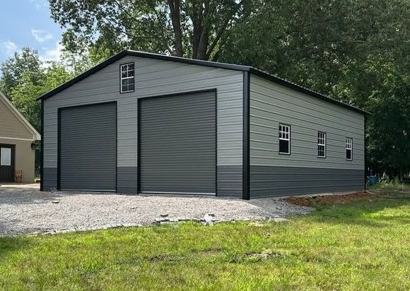 A two-tone grey metal garage with two roll-up doors, windows on the side, and a gravel driveway in a grassy yard.