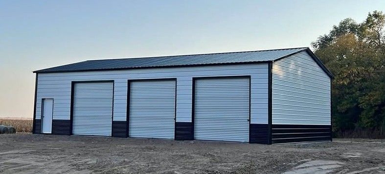 A white and black metal, three-bay garage building sits on a dirt lot with trees in the background.