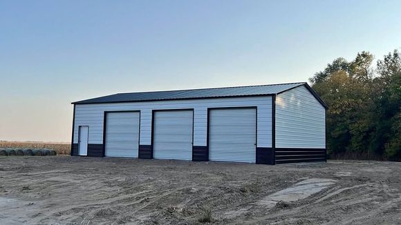 A white metal workshop with three roll-up doors and a side entry door, set on a dirt lot against a clear blue sky.
