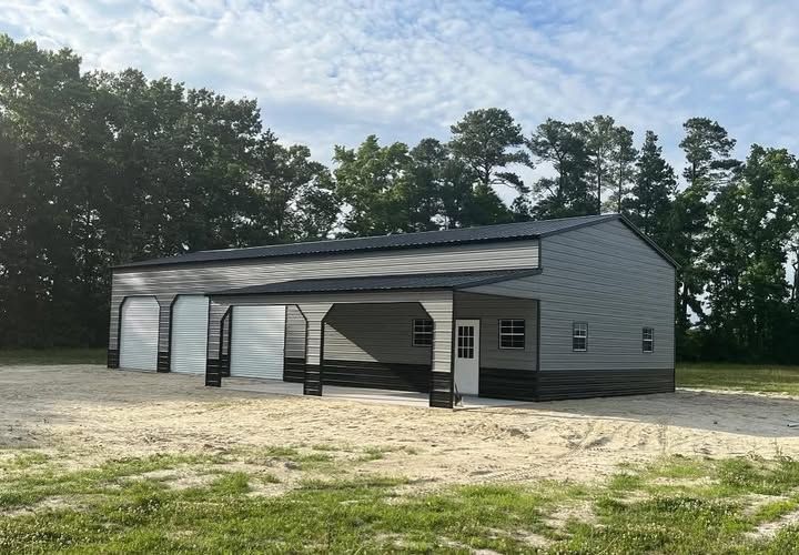 A gray and black metal building with three garage doors and an open-sided porch, set on a gravel lot near a treeline.