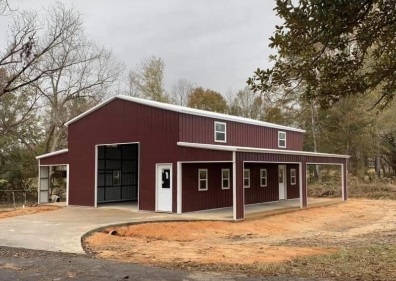 A maroon metal barn with a white trim, side lean-tos, and windows, set on a gravel lot near trees.