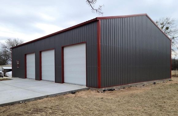 A dark metal-sided storage building with three white roll-up doors and red trim, set on a concrete slab in a grassy area.