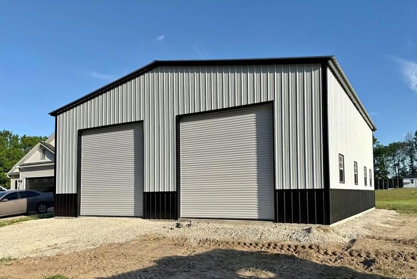 A modern, metal-sided barn with light gray panels, black trim, and two roll-up garage doors on a gravel lot.