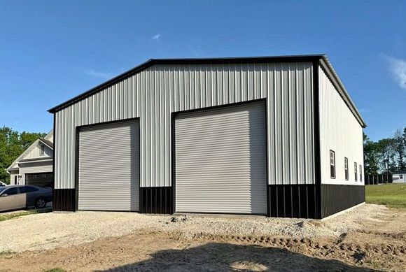A modern metal workshop building with light gray vertical panels, black trim, and two roll-up garage doors on a gravel lot.