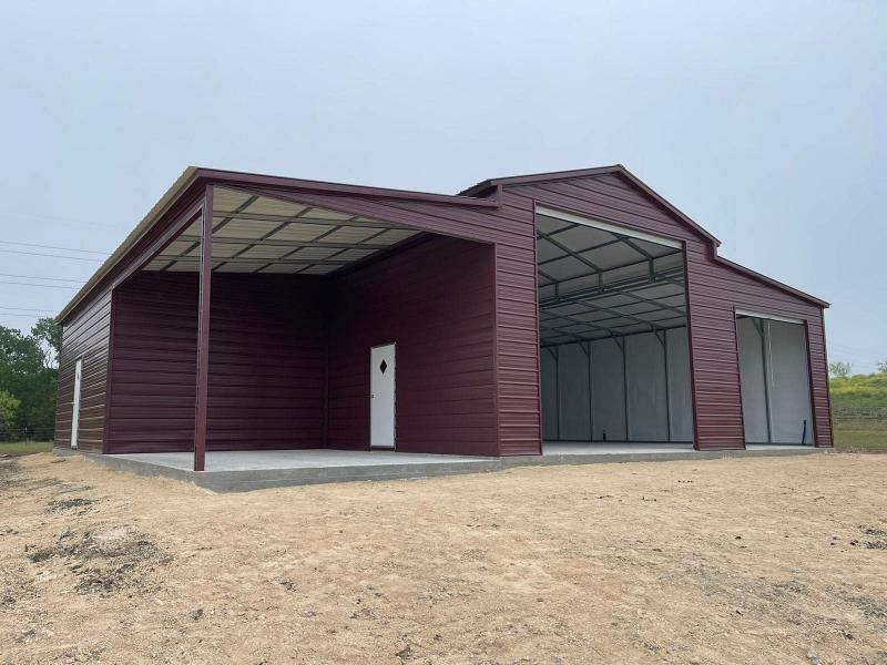 A maroon metal barn with a covered porch, large open bays, and white doors on a dirt lot under an overcast sky.