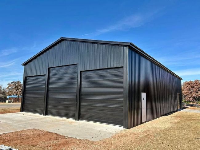 Black metal building with three garage doors against a blue sky. Concrete pad in front.