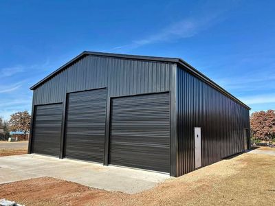 A modern, black metal workshop building with three roll-up doors, set on a concrete slab under a clear blue sky.