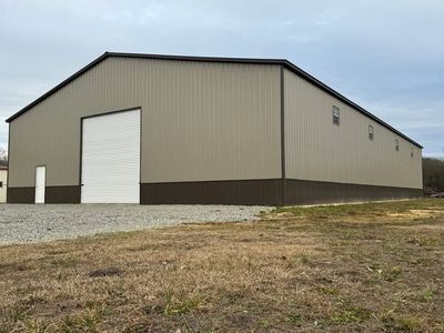 A large, beige metal warehouse with brown wainscoting, a large roll-up door, and a single pedestrian door on a gravel lot.