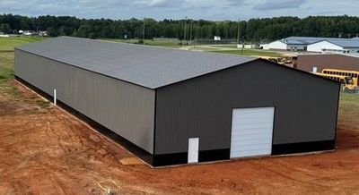 An aerial view of a long, gray metal building with a white door and garage bay, set on a reddish-brown dirt lot.