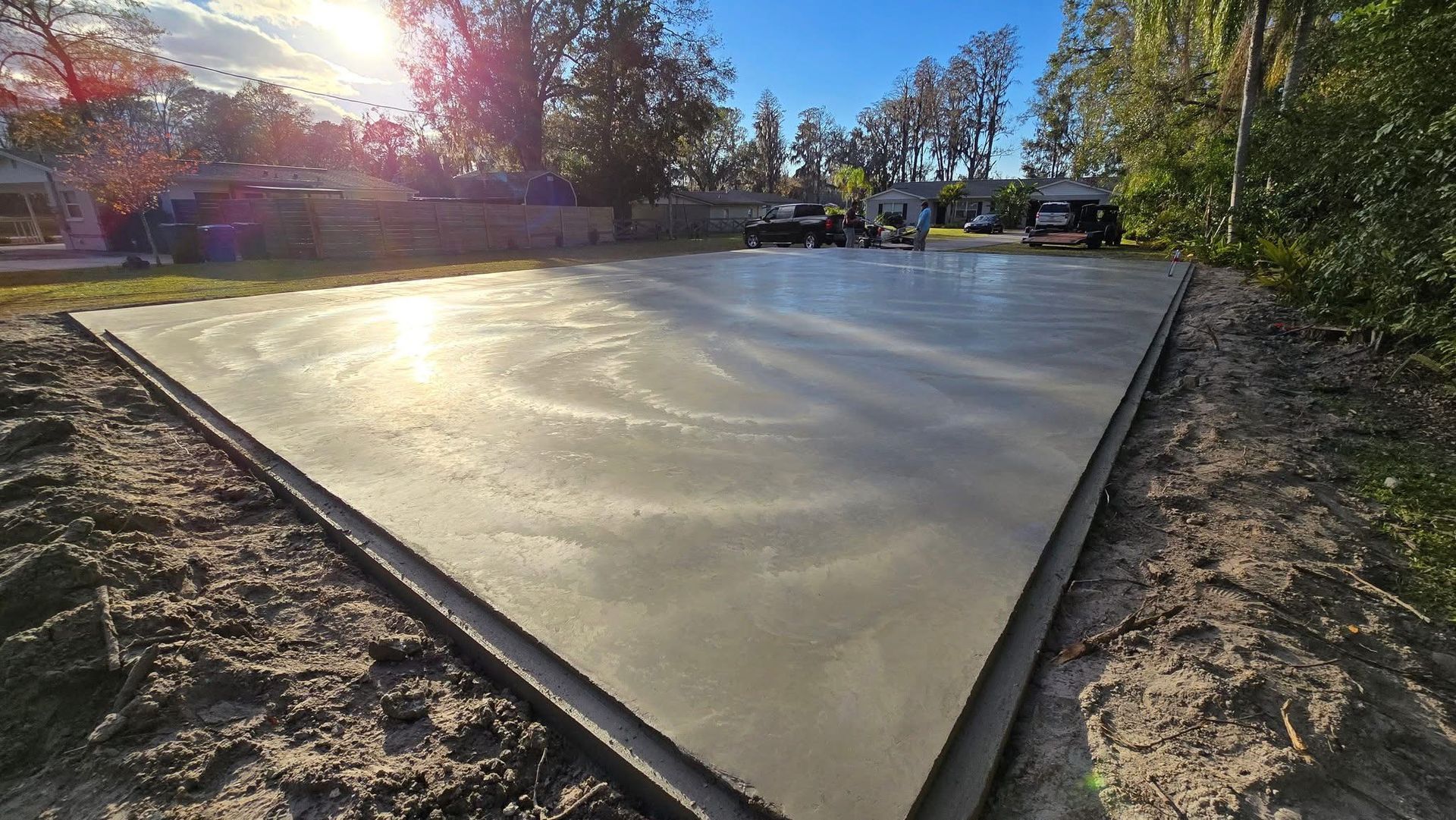 A freshly poured concrete slab foundation sits in a yard under bright sunlight, surrounded by dirt and trees.
