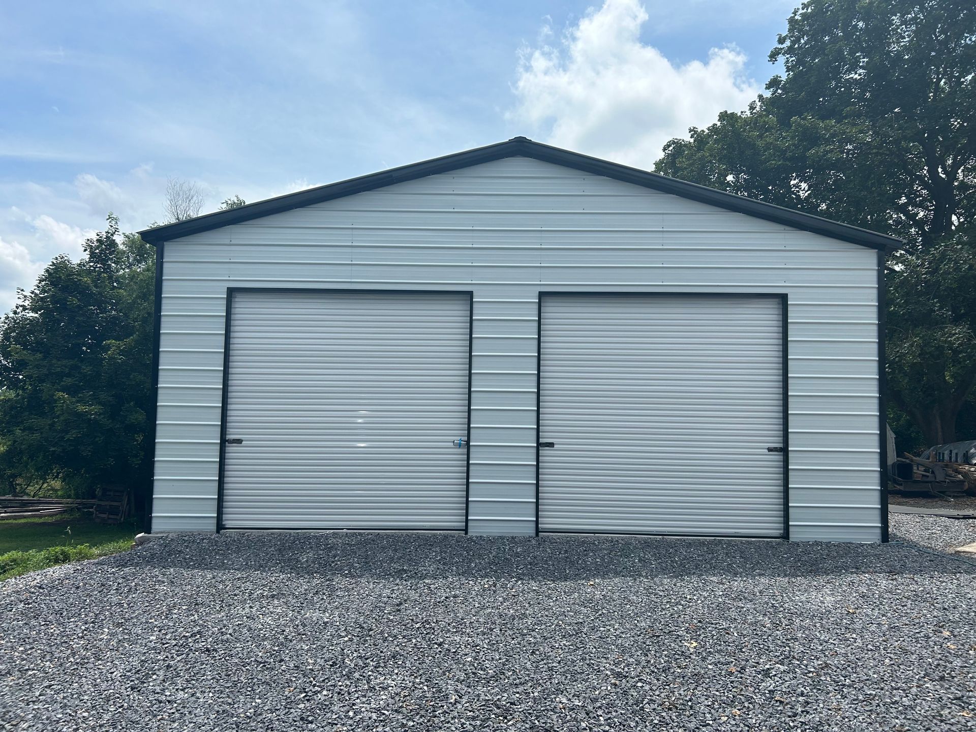 A light gray metal two-car garage with black trim stands on a gravel driveway under a partly cloudy sky.