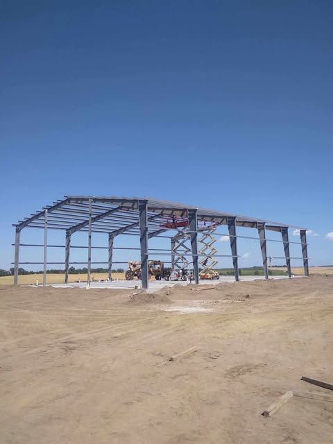 A metal building frame under construction on a dirt lot under a clear blue sky.