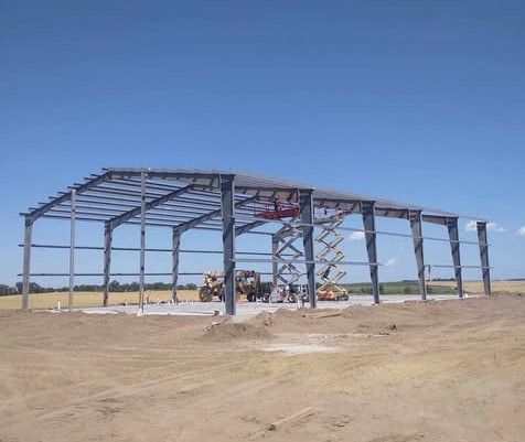 Steel frame of a building under construction on a concrete foundation in a field under a blue sky.