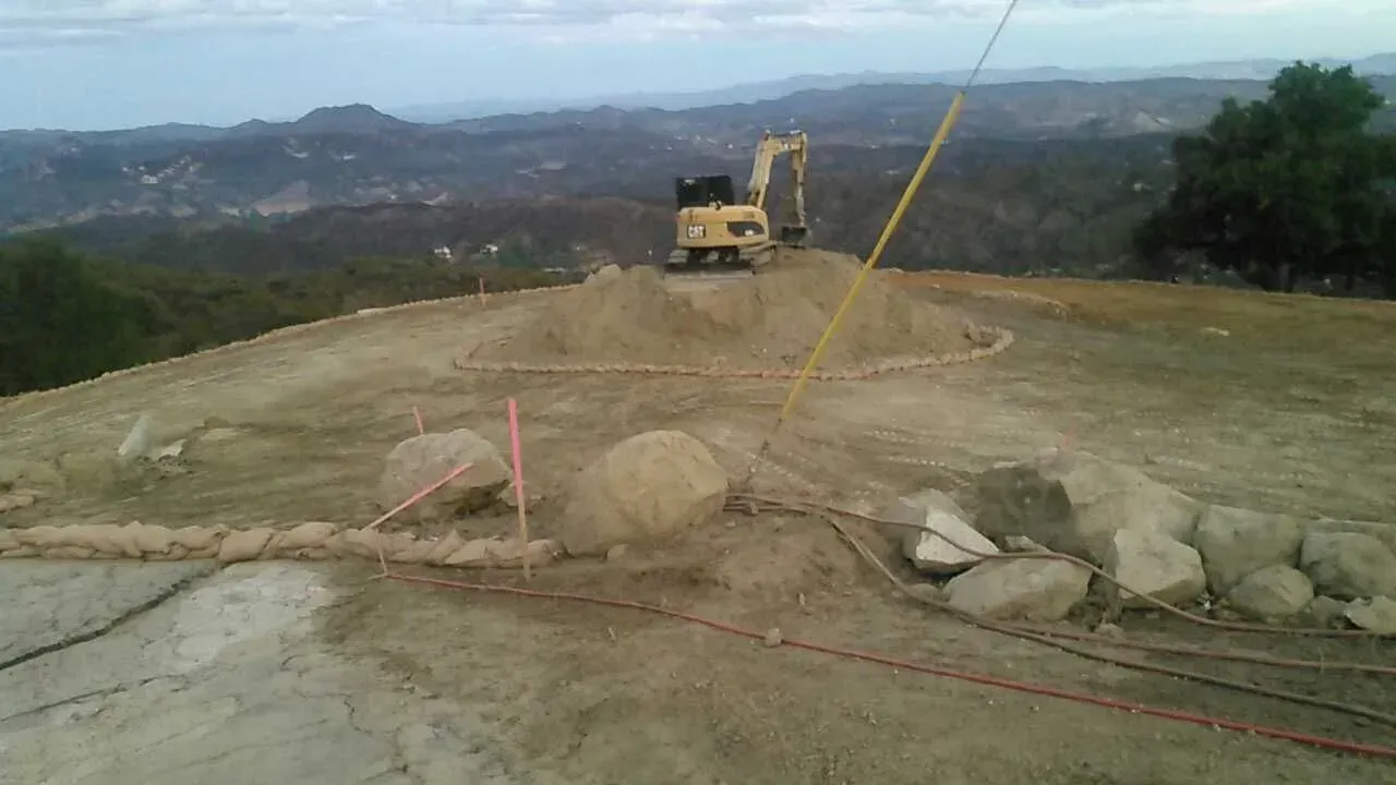 Construction site with excavator on a hilltop, mountains in background. Earthmoving and rock placement in progress.