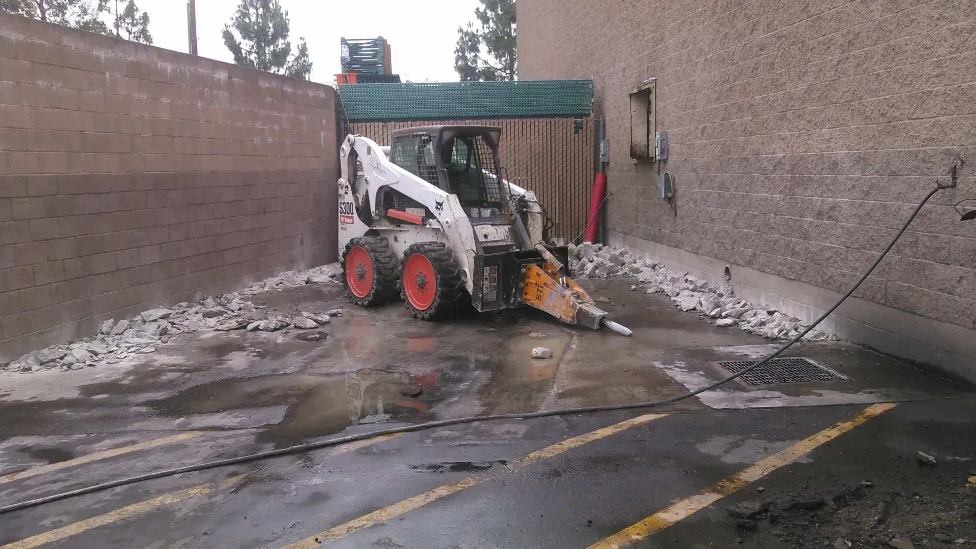Bobcat with jackhammer demolishes building wall in a parking lot.