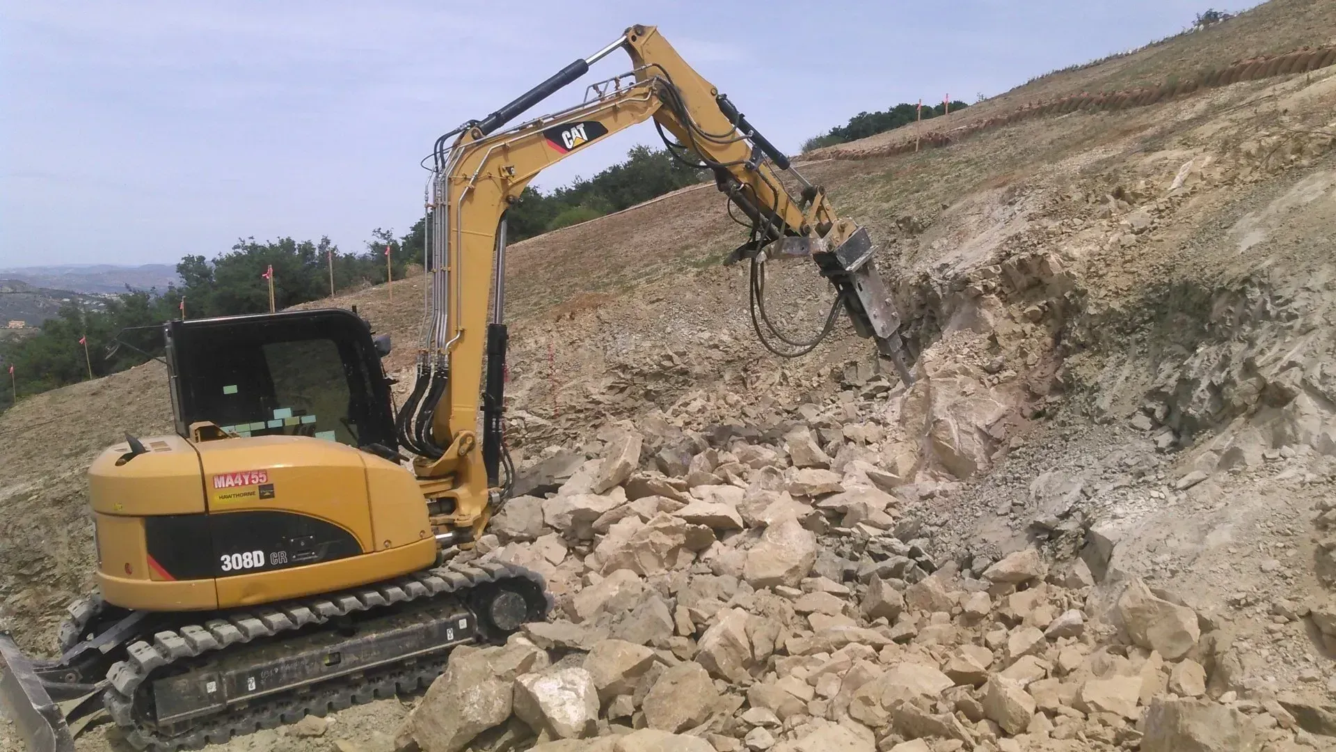Yellow excavator with a jackhammer breaking rocks on a hillside.