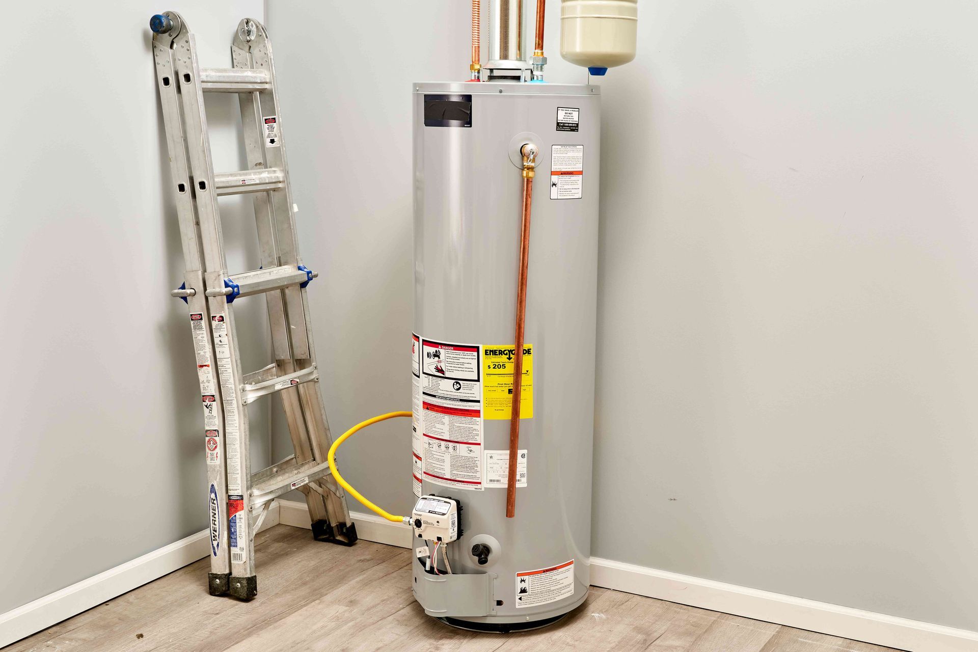 Water heater with ladder in corner of a room, gray tank with copper pipe and yellow electrical wire.