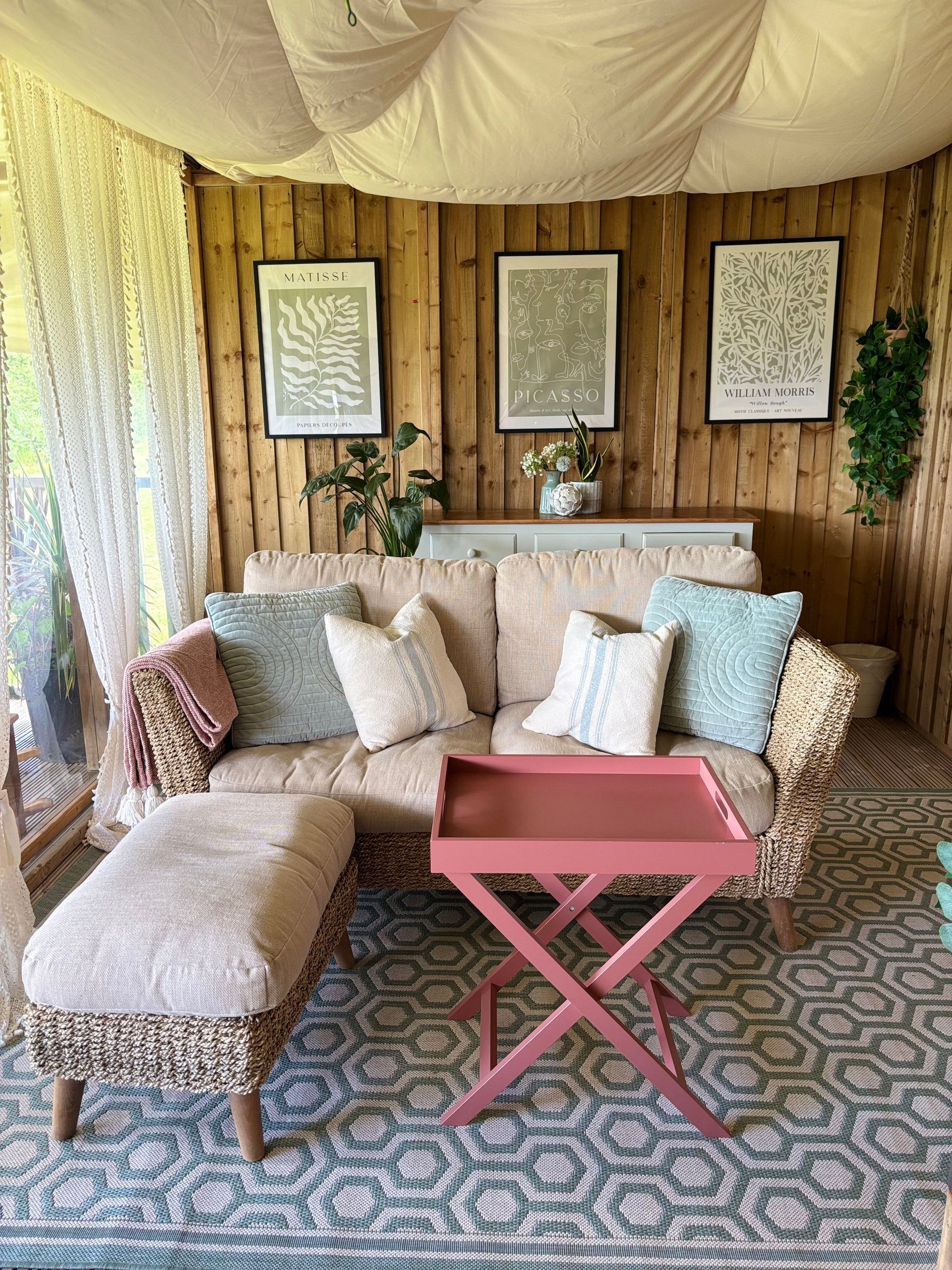 Cozy enclosed outdoor seating area with a sofa, ottoman, tray table, and botanical art on the wooden wall.