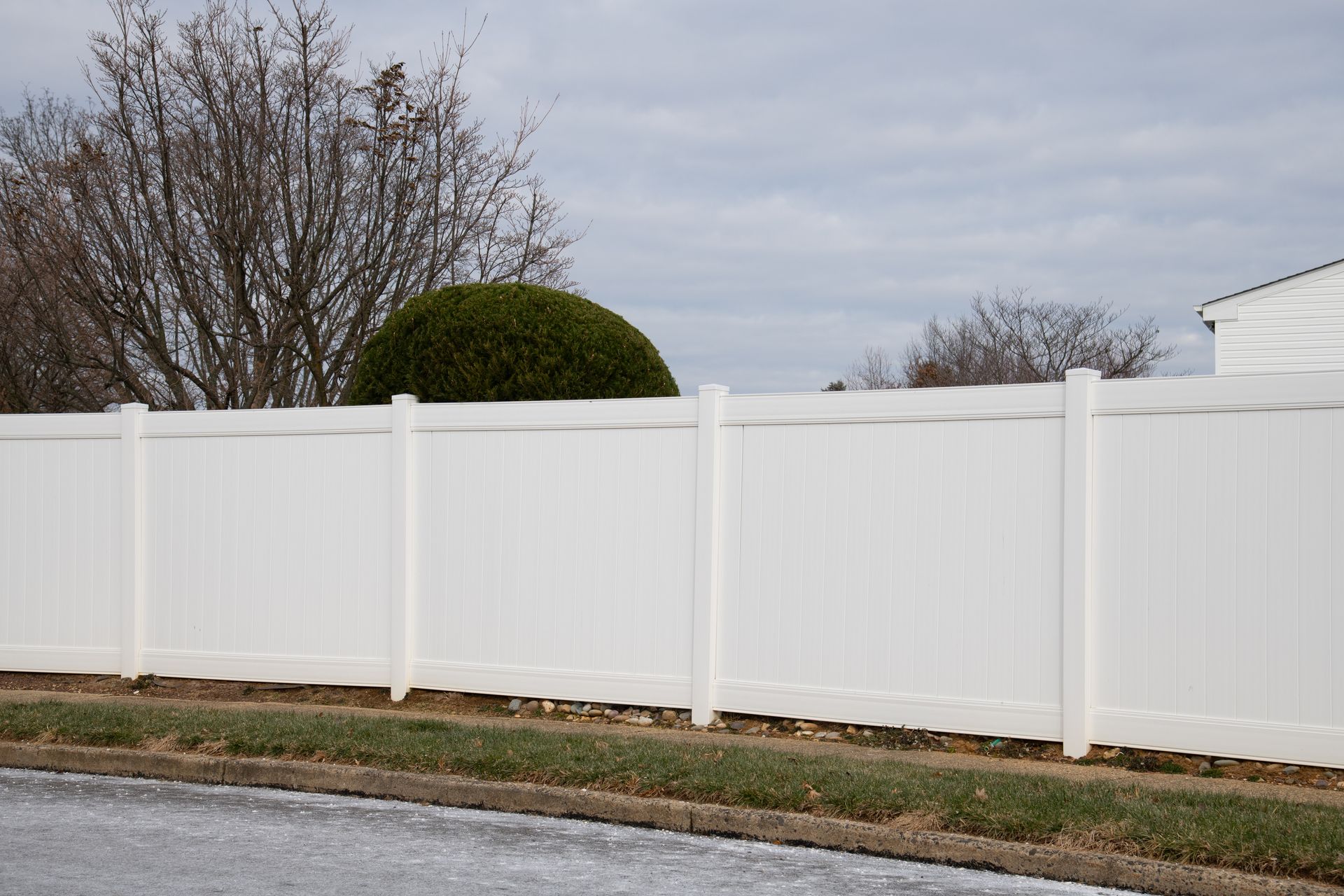 White vinyl fence along a road, with green shrubbery and bare trees visible behind it under a cloudy sky.