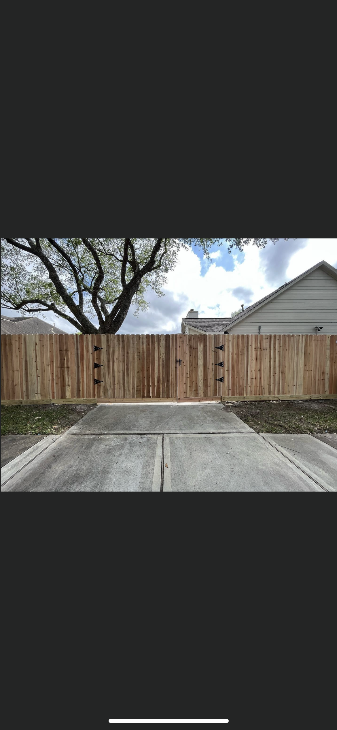 Wooden fence with a gate at the end of a concrete driveway, under a cloudy sky.