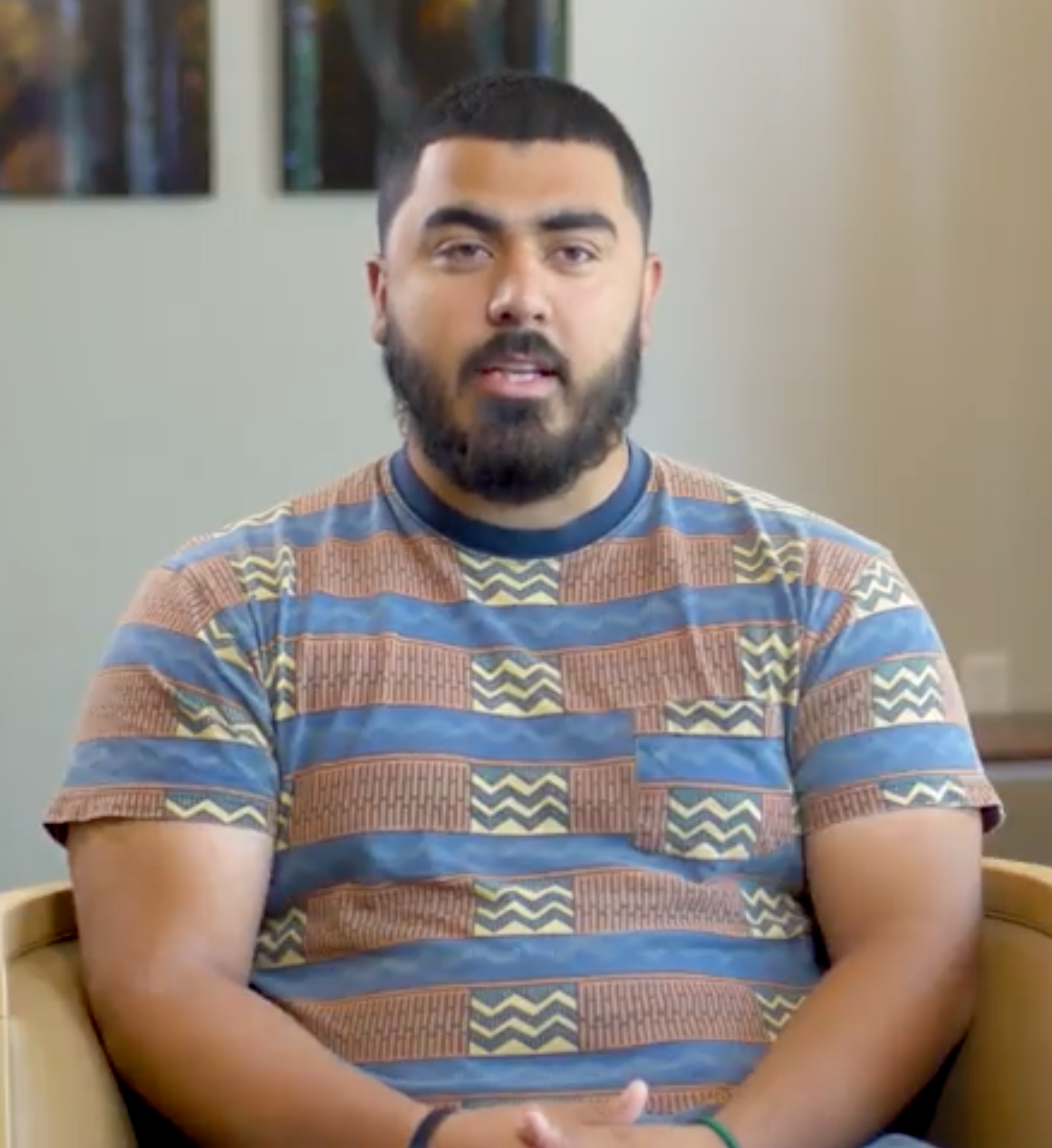 Man with a beard wearing a striped shirt sitting and talking. Indoors.