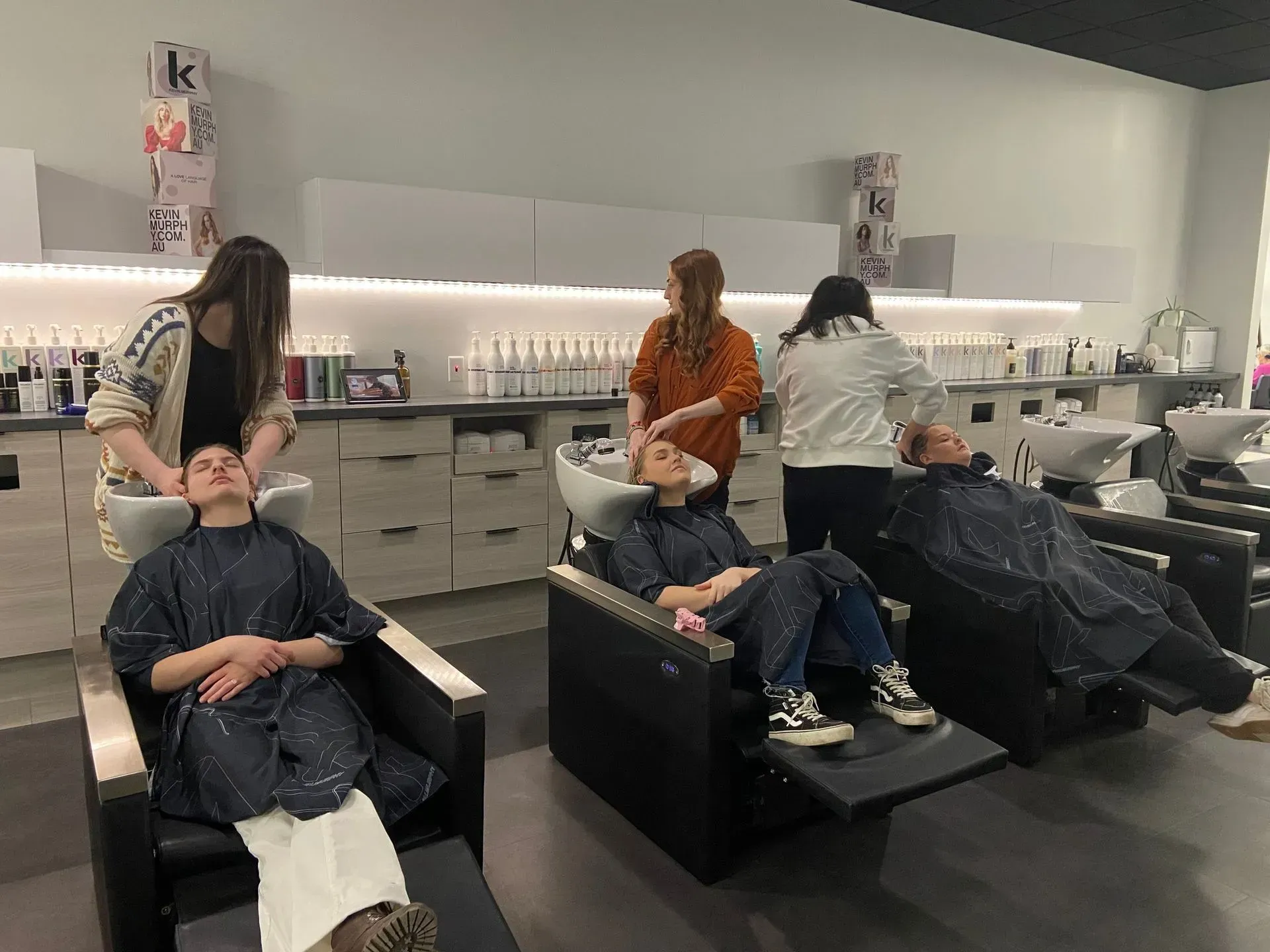 Salon customers getting hair washed by stylists in black chairs under bright lighting