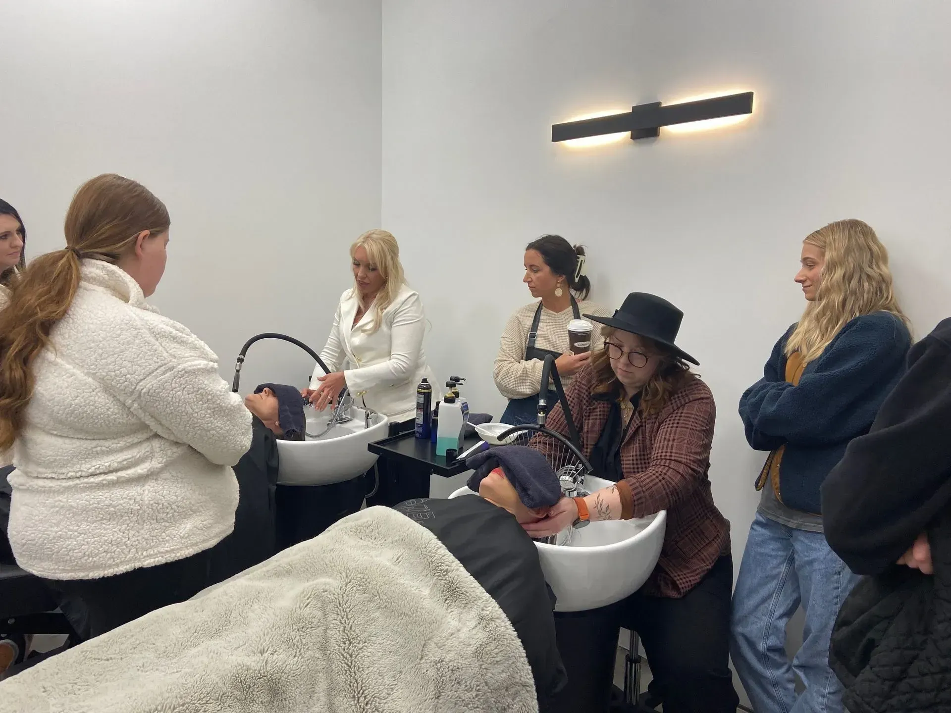 People in a salon washing hair in white basins, with a stylist serving clients in a modern room.