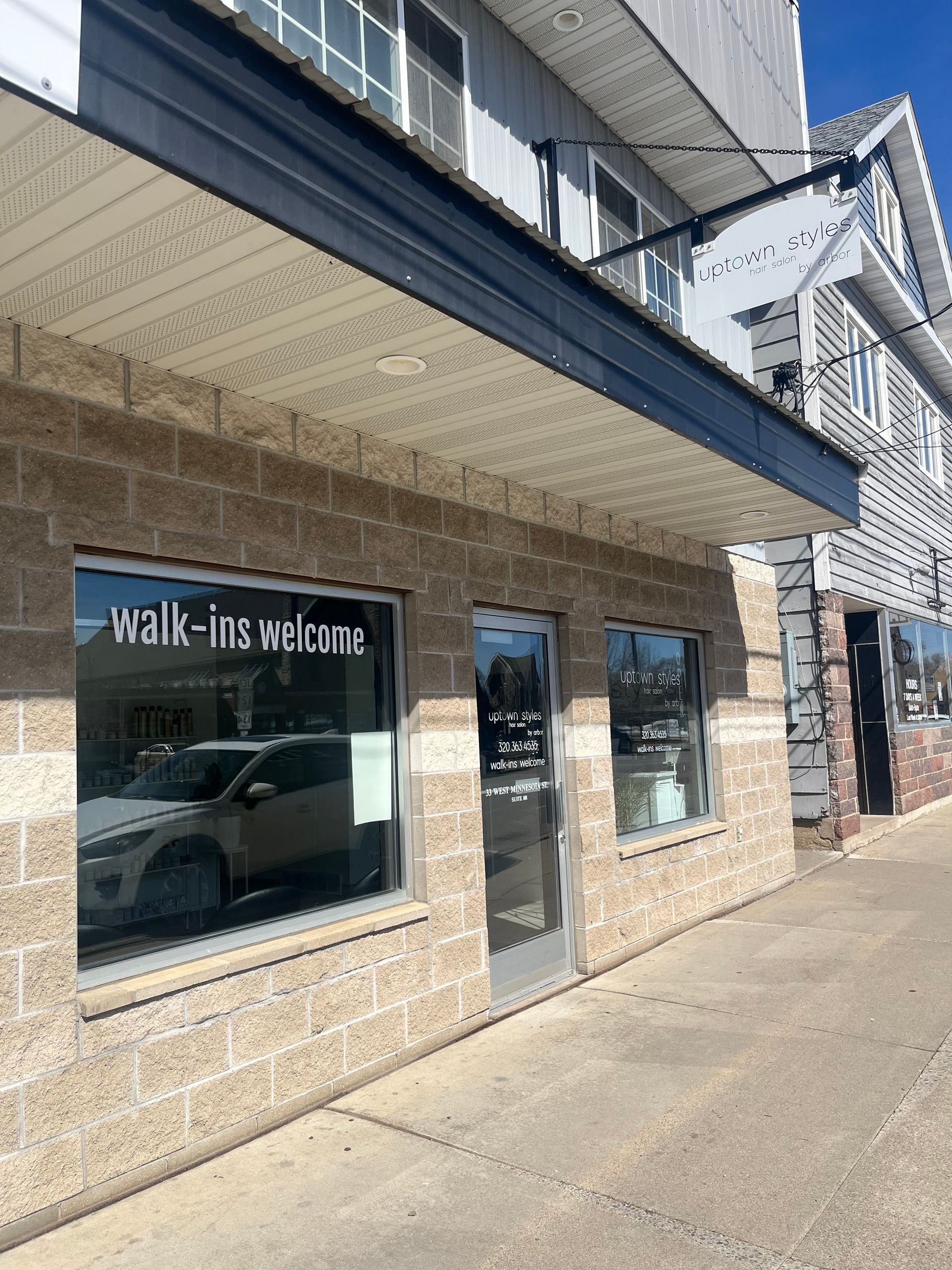 Storefront with “walk-in welcome” sign on a sunny sidewalk, with large windows and stone columns.