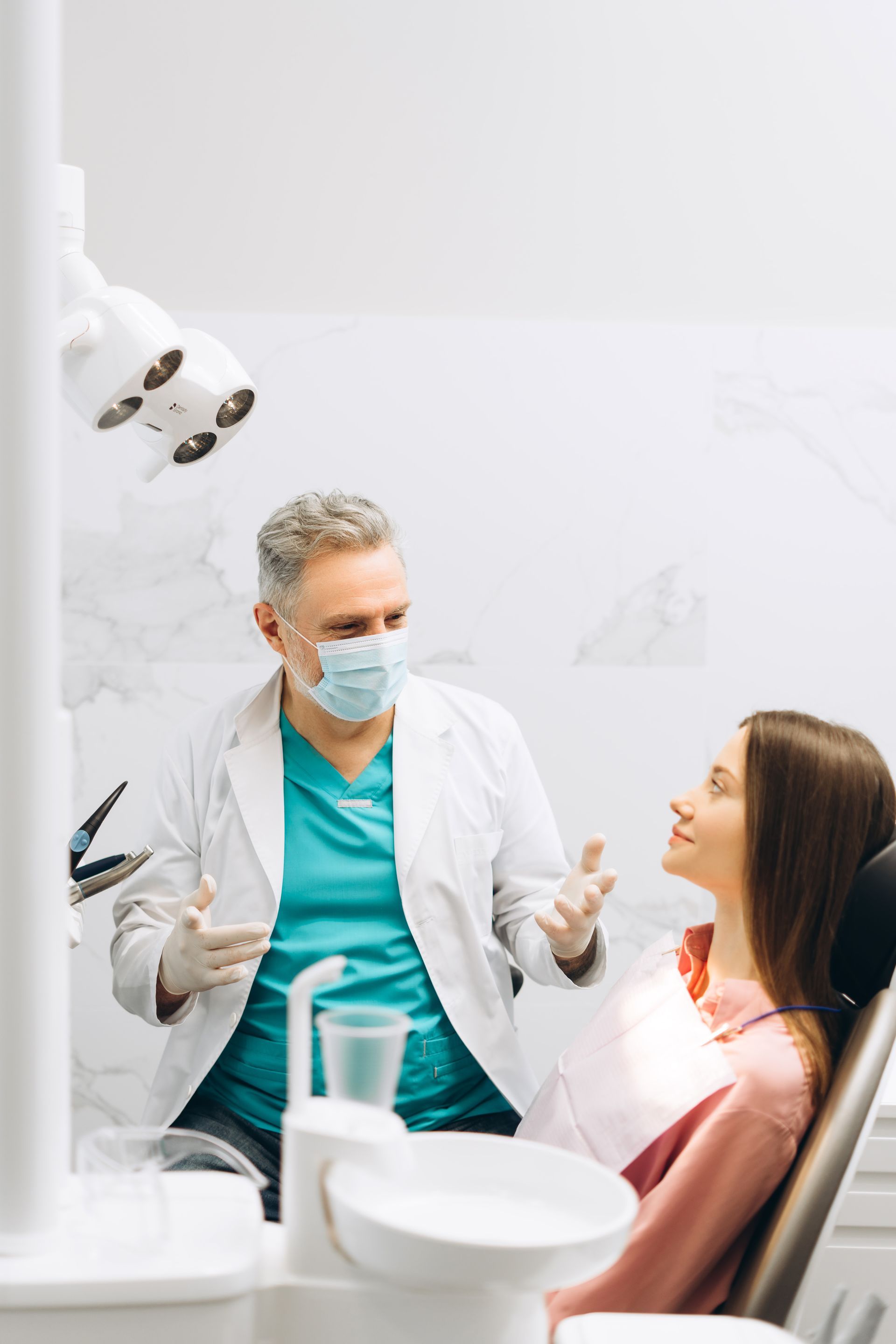Dentist speaking with a seated patient in a bright dental clinic, wearing masks and teal scrubs.