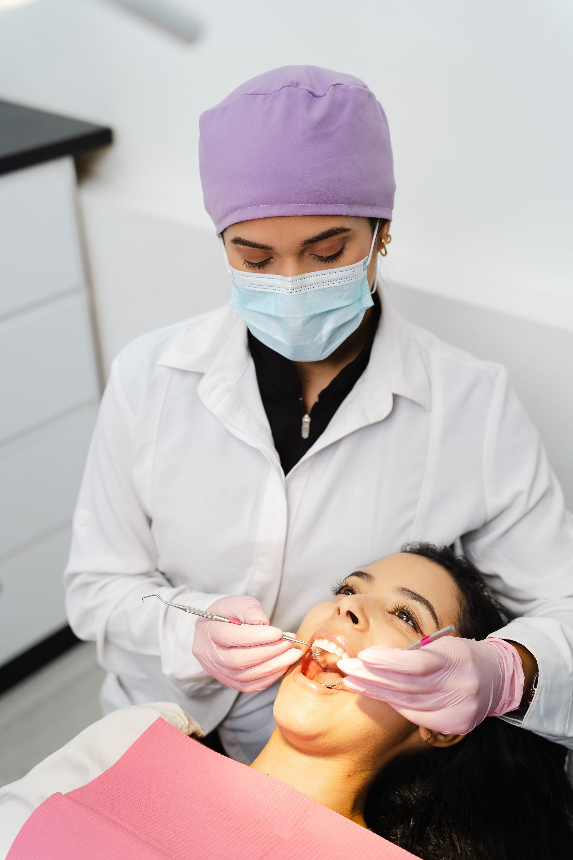 Dentist in purple cap and mask examining a patient’s mouth in a clinic