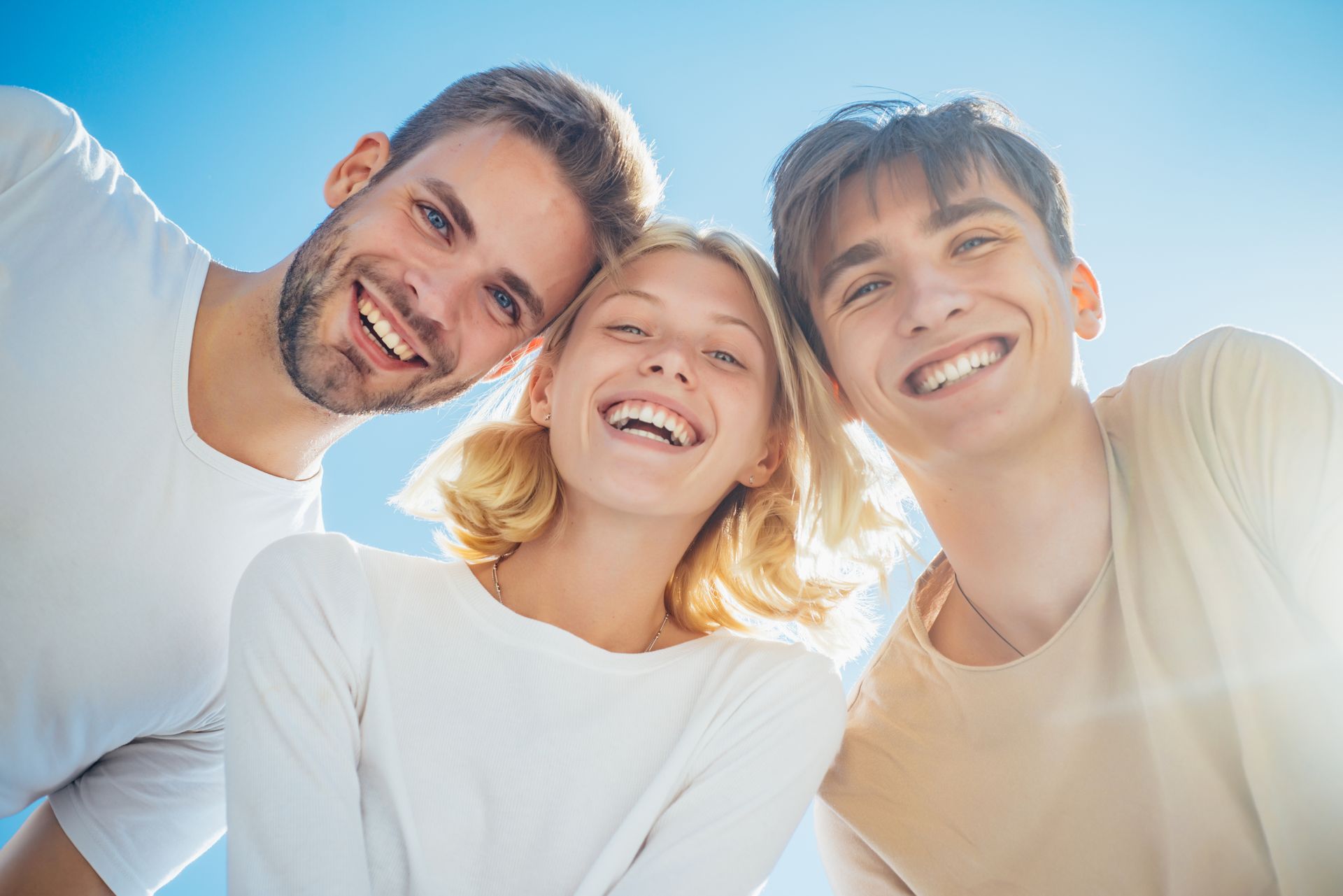 Three smiling people leaning close together outdoors in bright sunlight