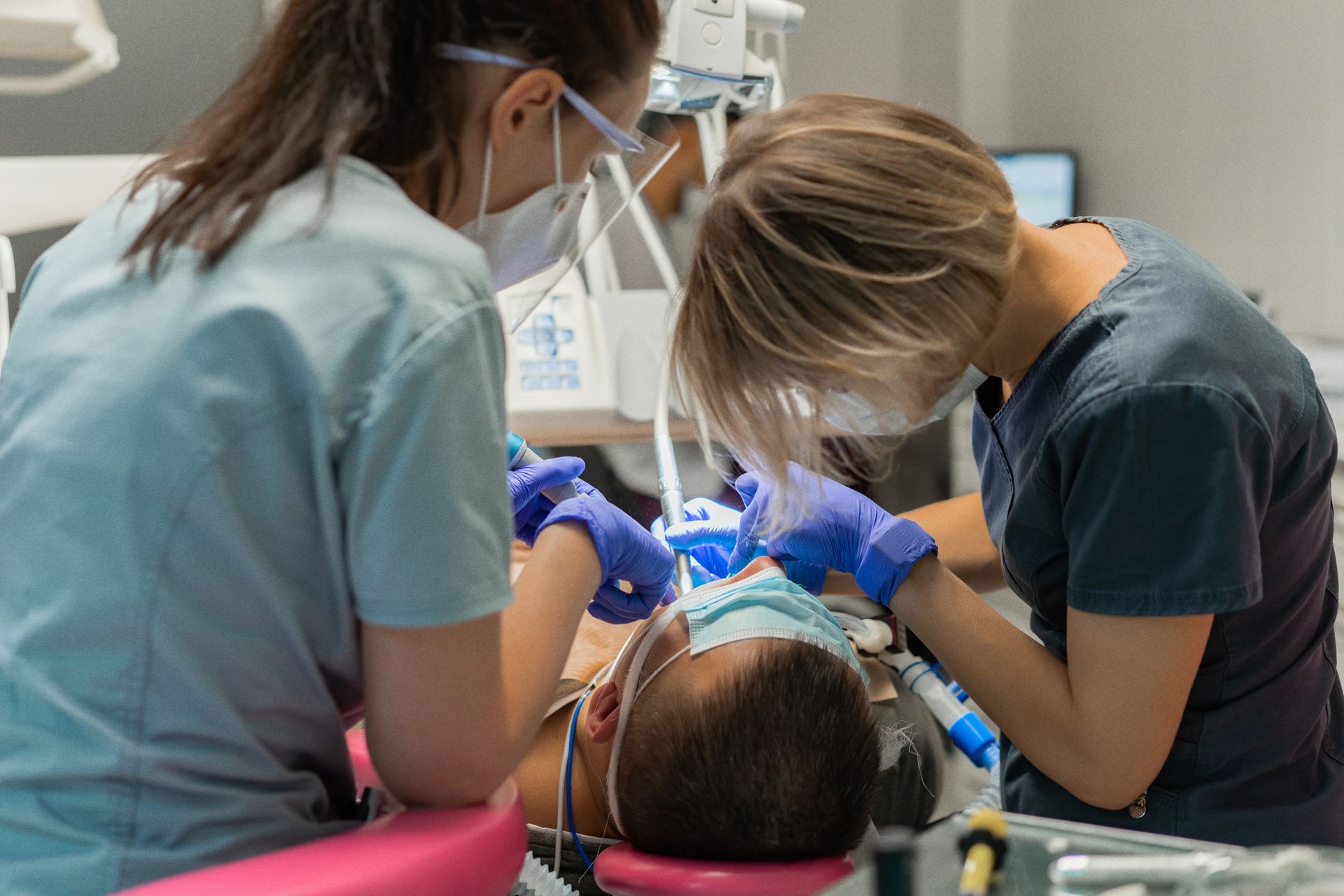Dental staff treating a patient in a clinic, wearing masks and blue gloves.