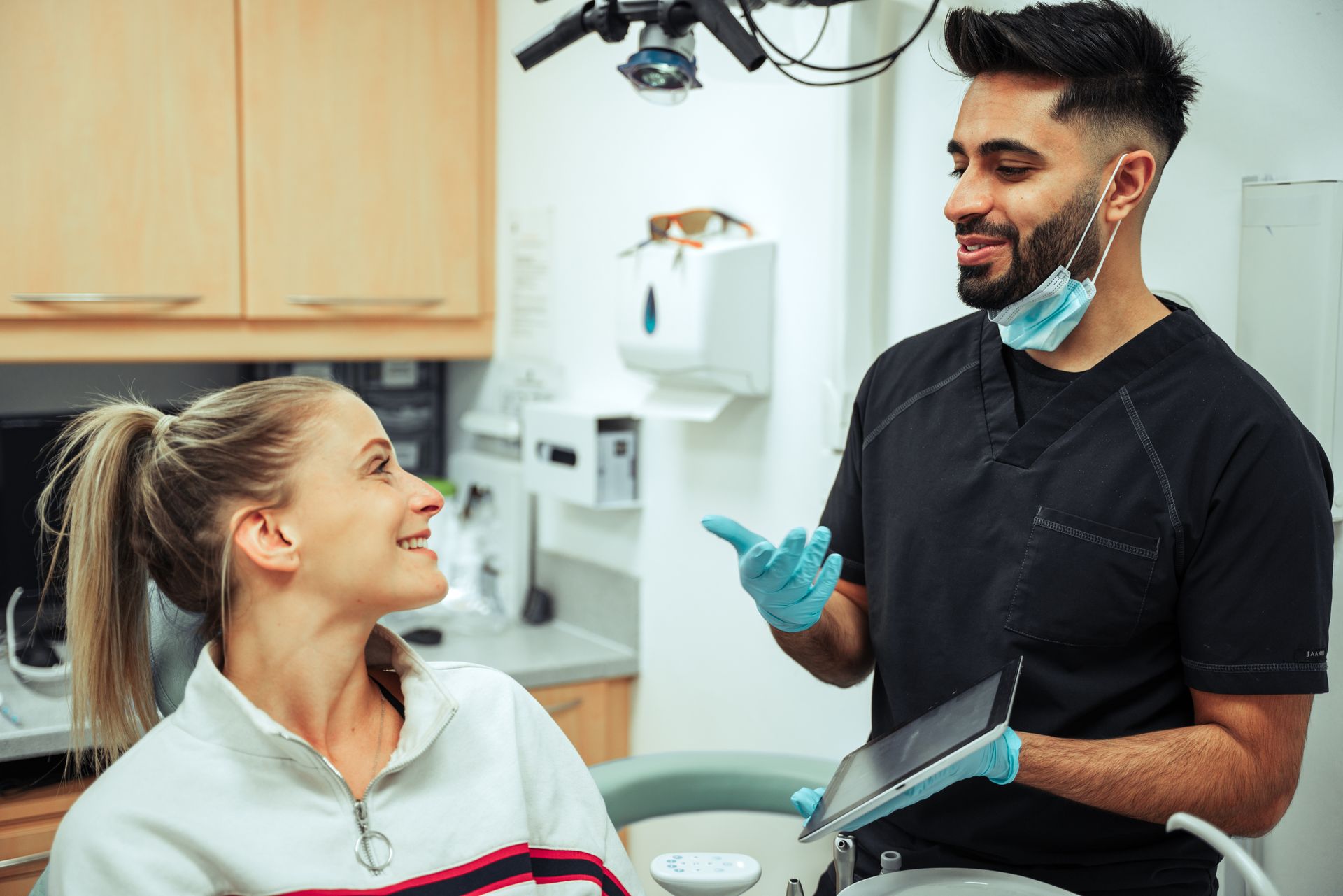 Dentist talking with a patient in a dental office, holding a tablet and wearing blue gloves.
