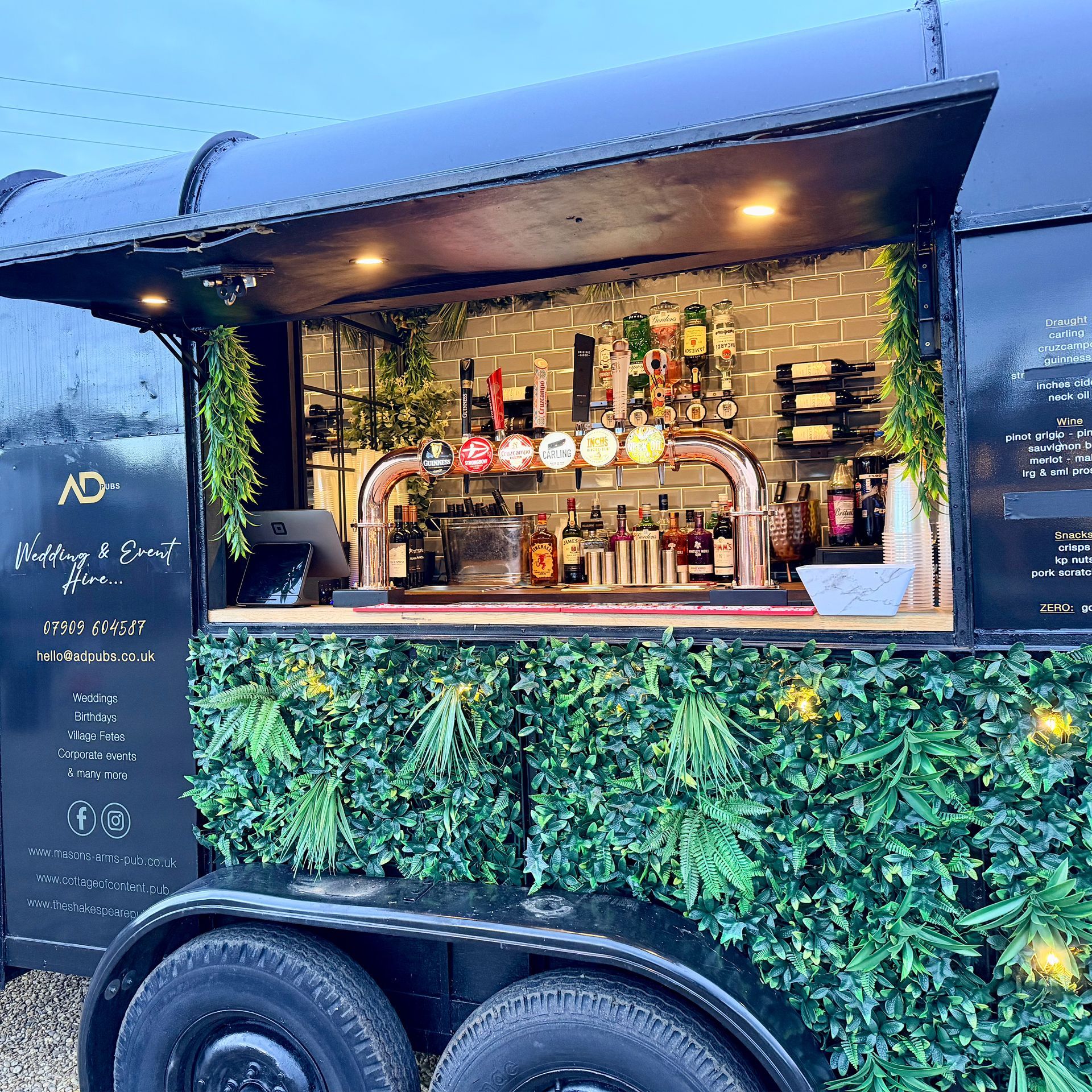 Black mobile bar trailer with open window, decorated with greenery and featuring taps and bottles.