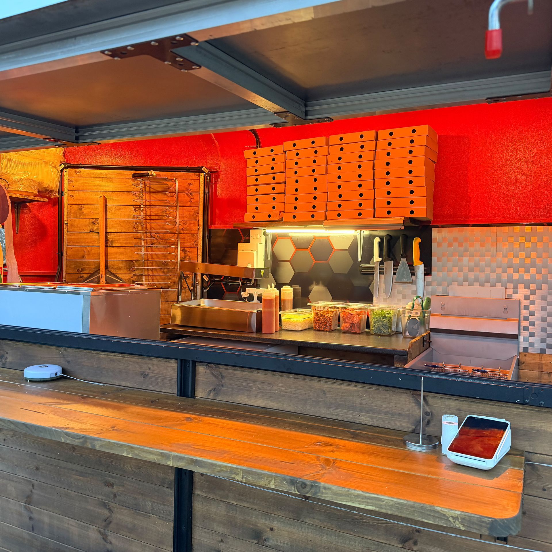 A food stand with a wooden countertop. The back wall is orange with an order menu, and there is a payment terminal on the counter.