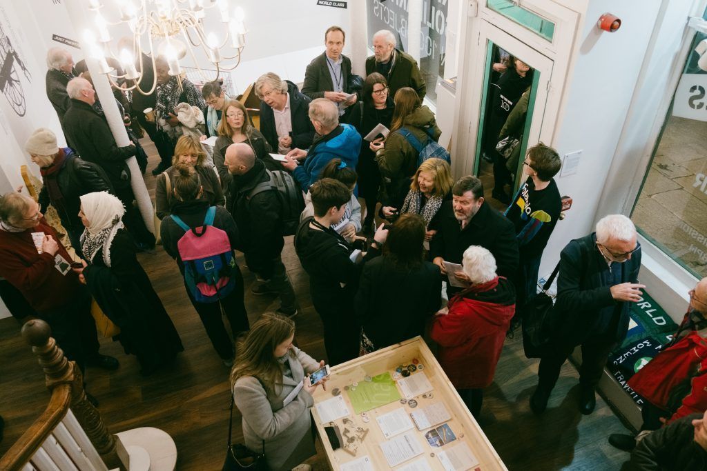 Crowded indoor gathering with people mingling around a table in a bright hallway