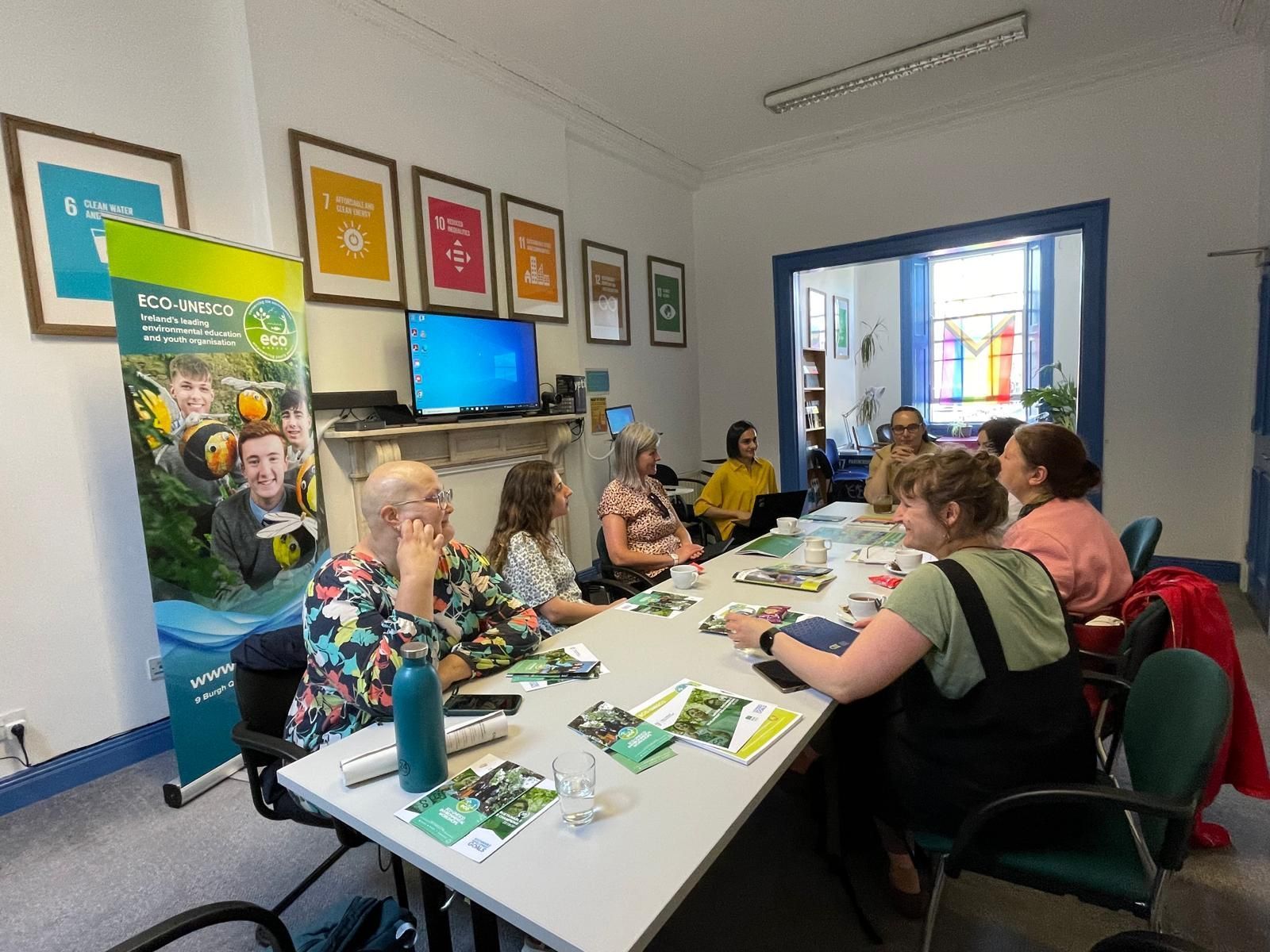 People seated at tables in a meeting room, with posters on the walls and a screen at the front.