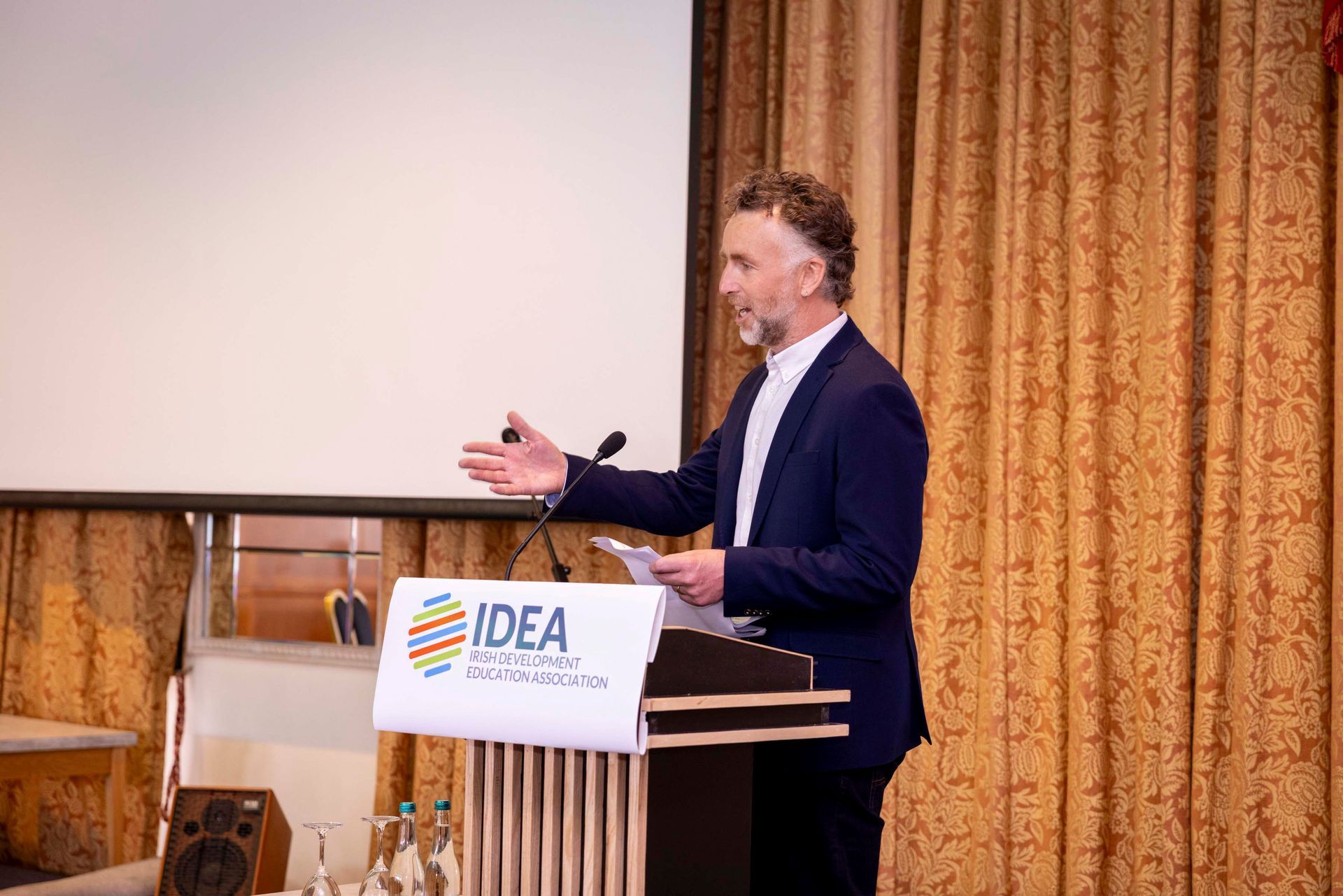 Man speaking at a podium with an IDEA sign, gesturing beside a projector screen and gold curtain backdrop