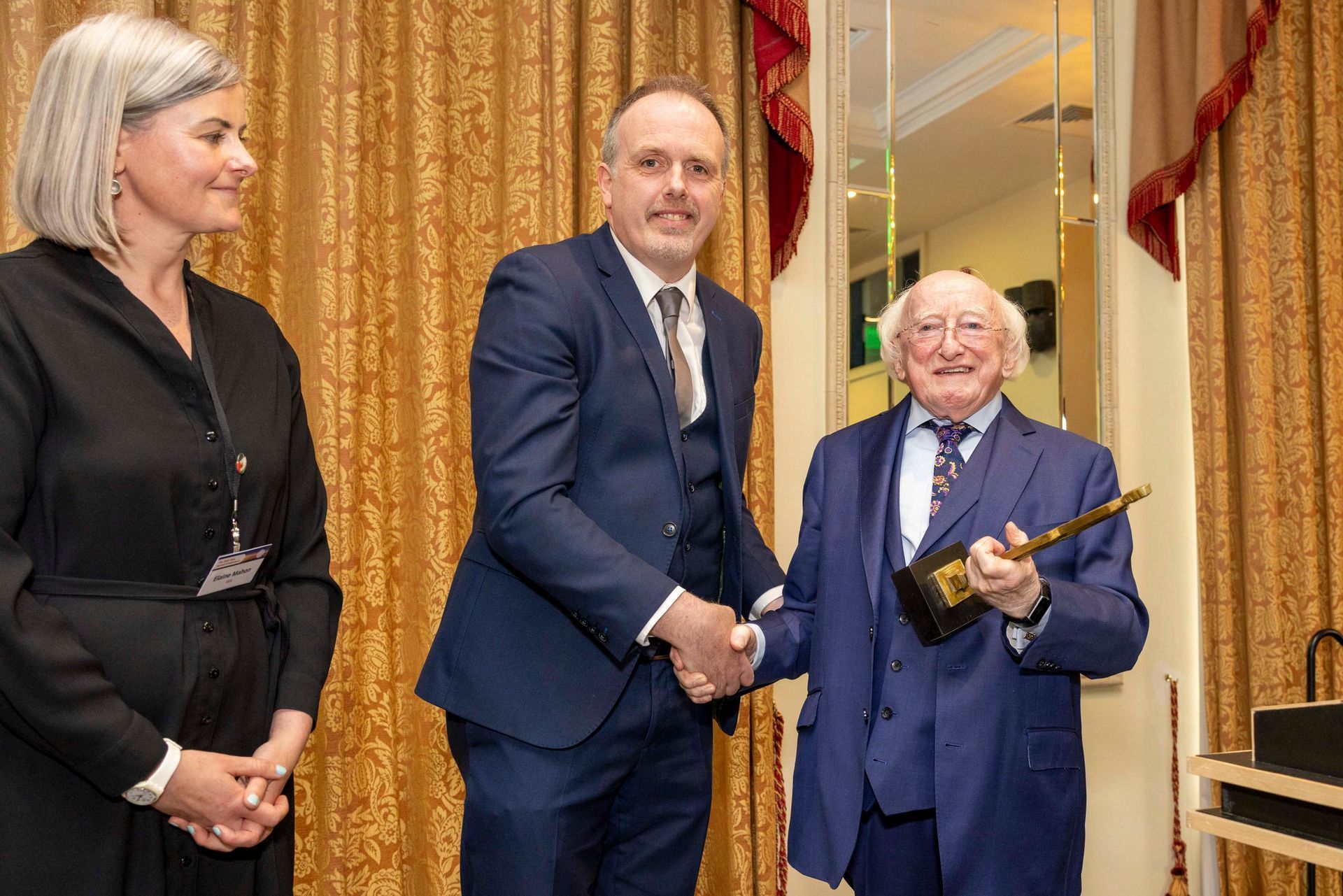 Three people at a formal ceremony, with two shaking hands while one holds a gold-topped object.