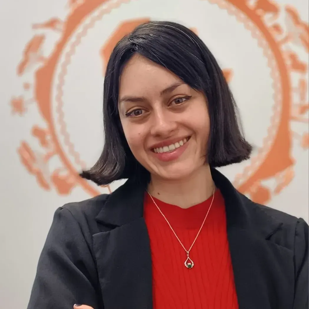 Smiling person in a black blazer and red top against a white wall with an orange circular mural