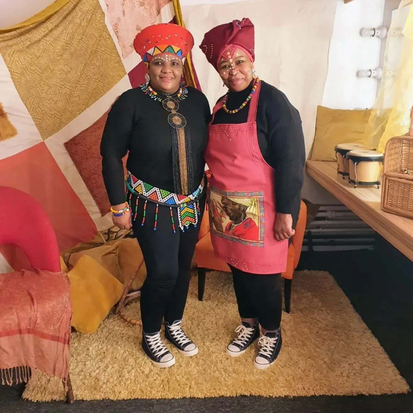 Two people in traditional red and black attire standing in a decorated room, smiling at the camera.