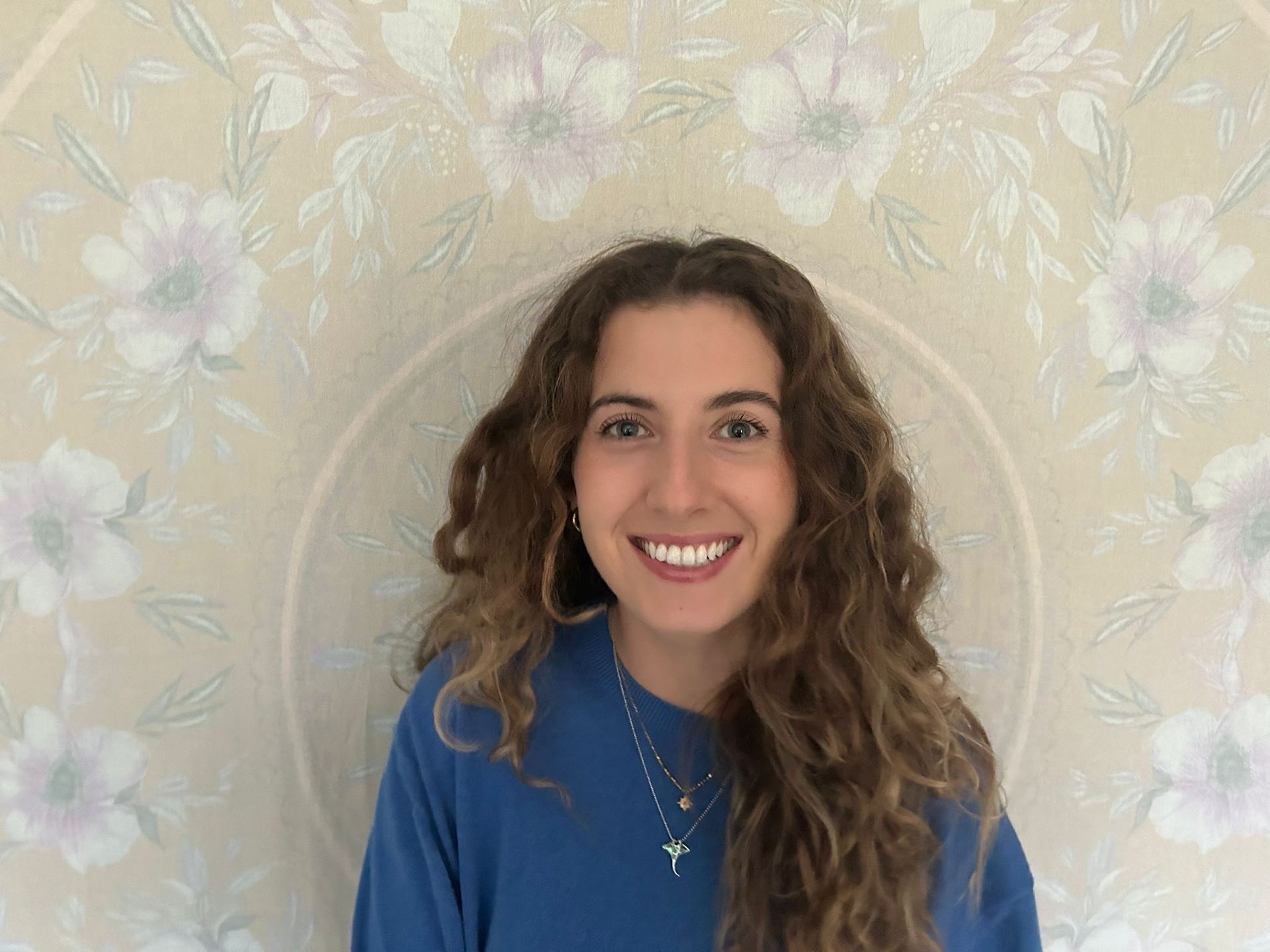 Smiling person in a blue sweater against a floral wallpaper background