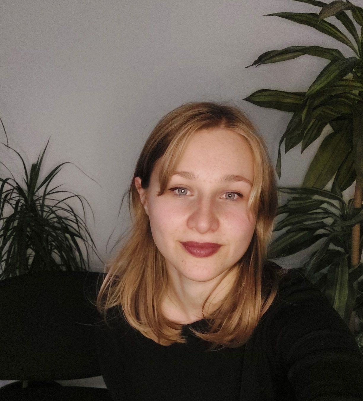 Portrait of a woman with shoulder-length hair, wearing a black top, seated indoors with houseplants behind her